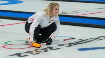 Jennifer Jones est accroupie sur la glace, regardant son lancer de pierre. Ses pieds sont sur la glace, juste au-dessus du logo de Beijing 2022.