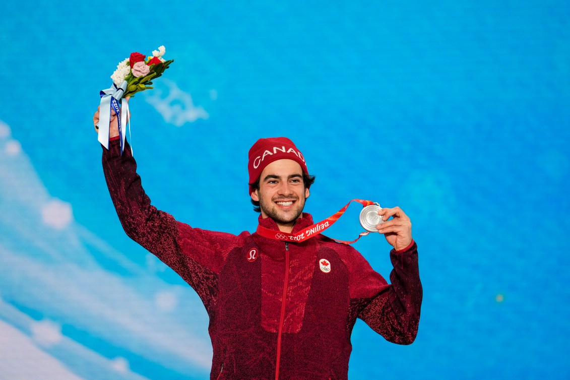 Le planchiste d'Équipe Canada Eliot Grondin soulève sa médaille d'argent avec sa main gauche et son bouquet de fleurs avec sa main droite sur le podium du snowboard cross masculin à Beijing 2022.