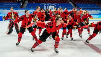 En avant-plan, Marie-Philip Poulin tient le drapeau canadien dans son dos comme une cape. En arrière-plan, le reste d'Équipe Canada célèbre la victoire en hockey féminin à Beijing 2022 avec sa médaille d'or autour du cou.