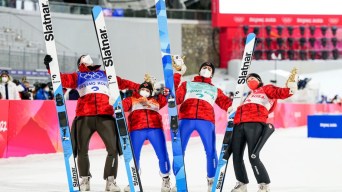 Les sauteurs à ski d'Équipe Canada Abigail Strate, Matthew Soukup, Alexandria Loutitt et Mackenzie Boyd-Clowes tiennent leurs skis de la main droite, la mascotte des Jeux de la main gauche et ils se penchent vers l'arrière pour célébrer leur médaille de bronze à l'épreuve de saut à ski mixte par équipes à Beijing 2022.