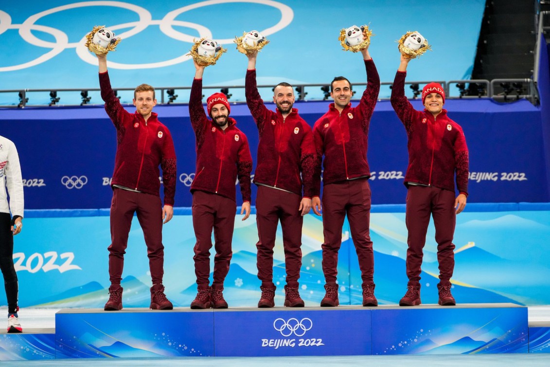Cinq patineurs sur le podium tiennent une figurine de la mascotte des Jeux en levant la main dans les airs.