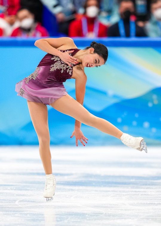 Madeline Schizas est sur la glace et se tient sur son patin droit. Elle est penché sur son côté, fixant patin droit relevé. Sa main droite est sur sa poitrine tandis que sa main gauche ouverte temd vers la glace à l'arrière de sa jambe relevée. Elle porte une robe chiffon courte mauve et rose avec détails en bijoux argent. Ses cheveux foncés sont lissés vers l'arrière, attaché avec une toque.