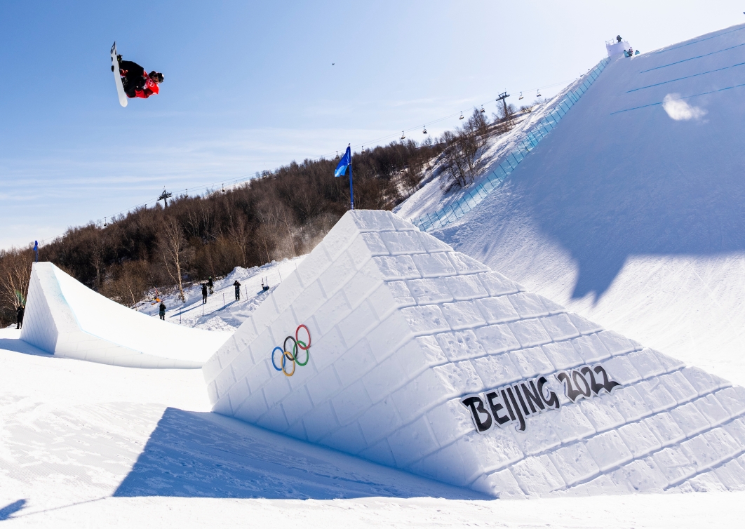 Le planchiste Mark Morris vêtu de rouge et noir est en plein saut dans les airs au milieu du parcours de l'épreuve du slopestyle, entre 2 pentes. On aperçoit la signature de Beijing 2022 en noir sur la neige d'une des pentes. Gros soleil et ciel bleu.
