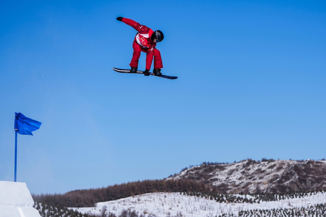 Le planchiste d'Équipe Canada Max Parrot est sur sa planche, en plain envol sur le fond du ciel bleu. Il a les deux pieds sur sa planche et la tien avec une main. Son autre bras est vers l'arrière. On voit au loin le terrain dénudé de neige avec un petit drapeau bleu en avant-plan gauche de l'image.