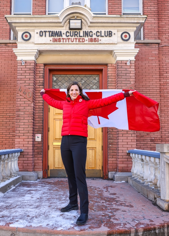 Lisa Weagle devant la porte du Club de curling tenant le drapeau canadien.