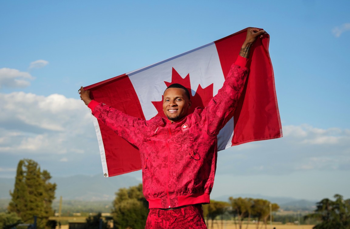 Le sprinteur Andre De Grasse pose habillé en rouge en tenant le drapeau canadien derrière lui.