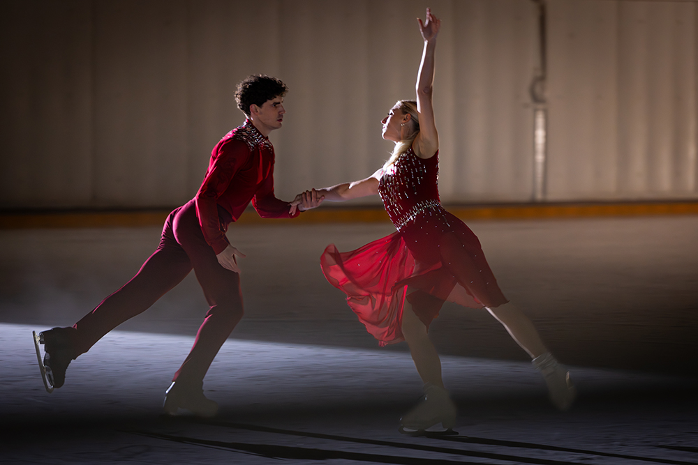 Piper Gilles et Paul Poirier sur une patinoire intérieure, vêtus de costumes rouges, patinant en se tenant la main.