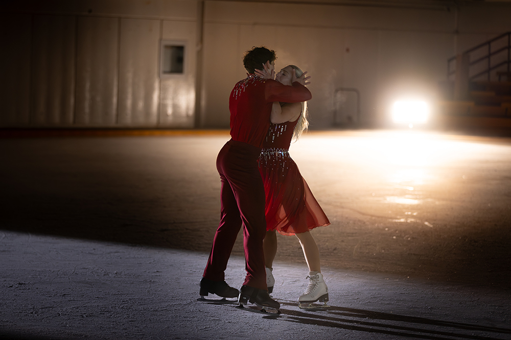 Piper Gilles et Paul Poirier, vêtus de costumes rouges, s'enlacent dans une patinoire intérieure.