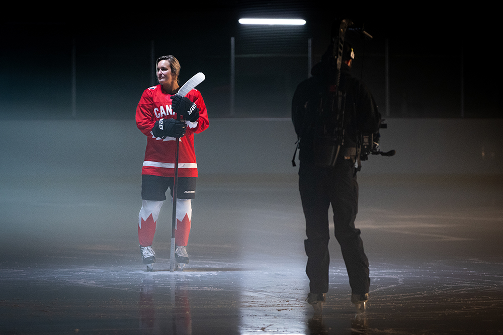 Marie-Philip Poulin dans un aréna intérieur, vêtue de l'équipement de hockey de l'équipe canadienne et d'un chandail rouge, debout, regardant de côté devant une caméra.