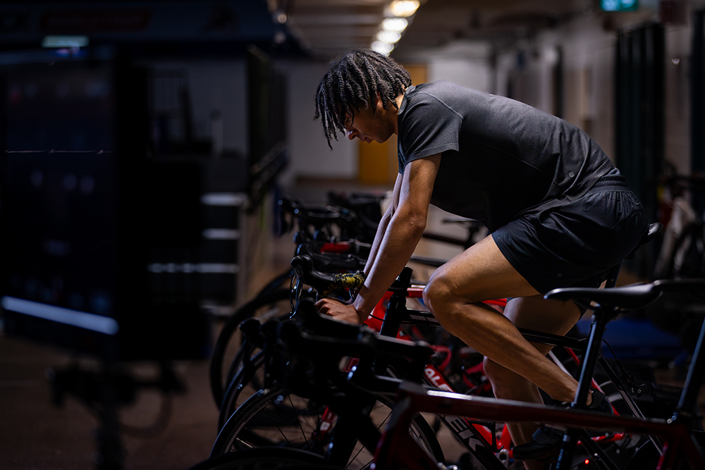 William Dandjinou, vêtu d'une tenue de sport noire, fait du vélo dans une pièce faiblement éclairée.