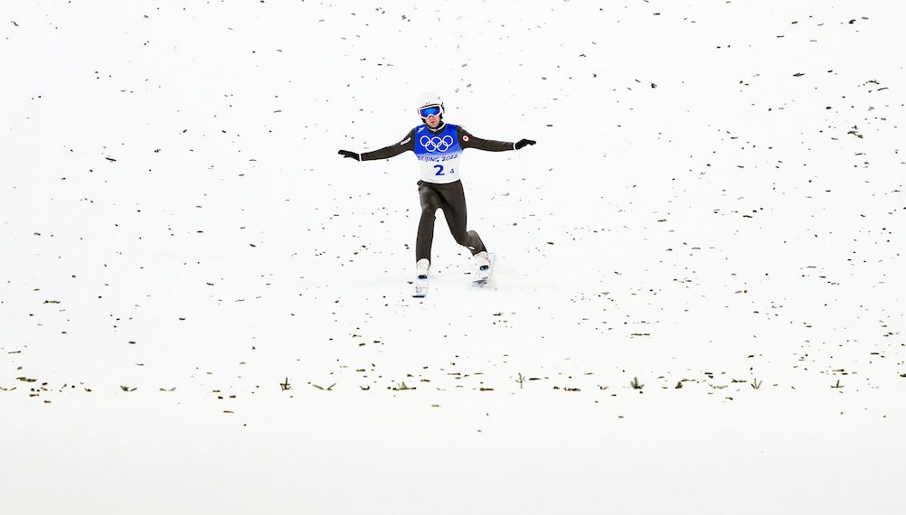 Ski jumper in black suit and blue bib shows a telemark landing on snow 