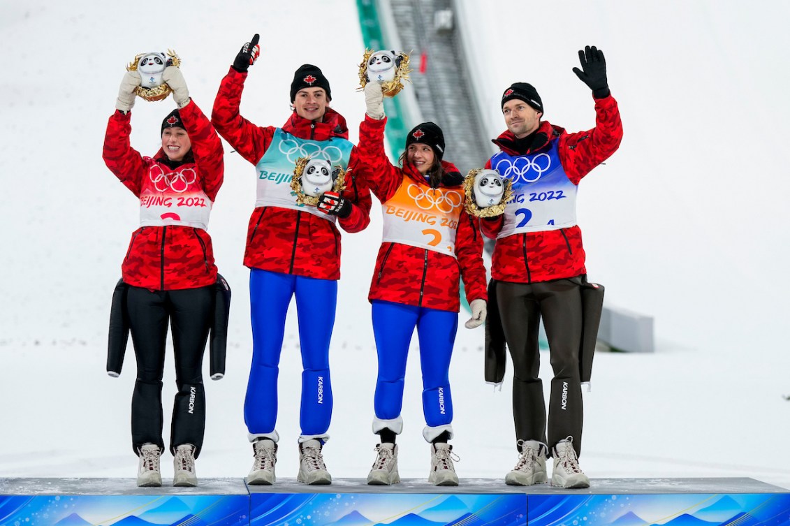 Four ski jumpers wearing red jackets hold up mascot stuffed animals on the podium 