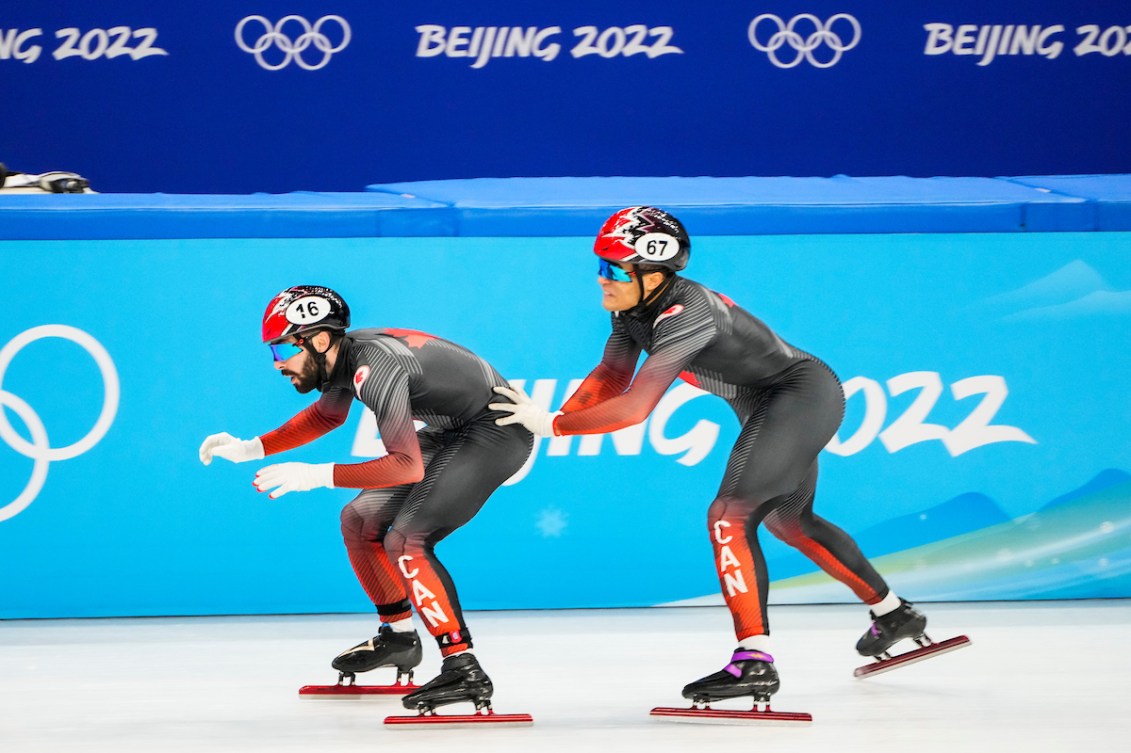 One Canadian short track speed skater pushes a teammate forward with hands on butt 