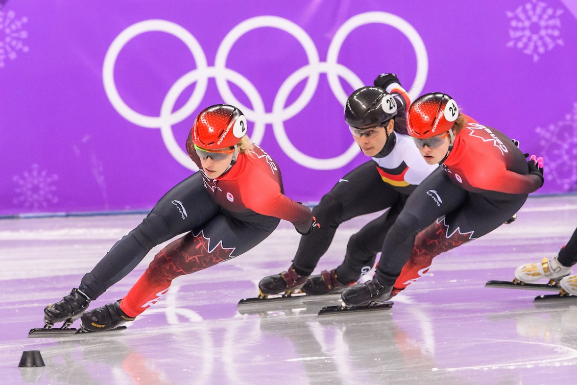 Two Canadian short track speed skaters race against a German opponent in front of a purple background 