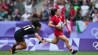 A Team Canada rugby player in a red jersey avoids a tackle from an opponent in a black jersey