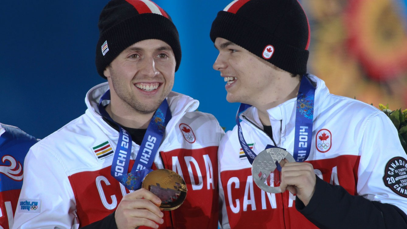 Mikael Kingsbury looks at Alex Bilodeau laughing who is standing at the Sochi 2014 medal presentation. Alex Bilodeau is smiling and holding his bronze medal and Kingsbury is holding him silver medal. They are dressed in their red and white Team Canada sweaters and toques. 