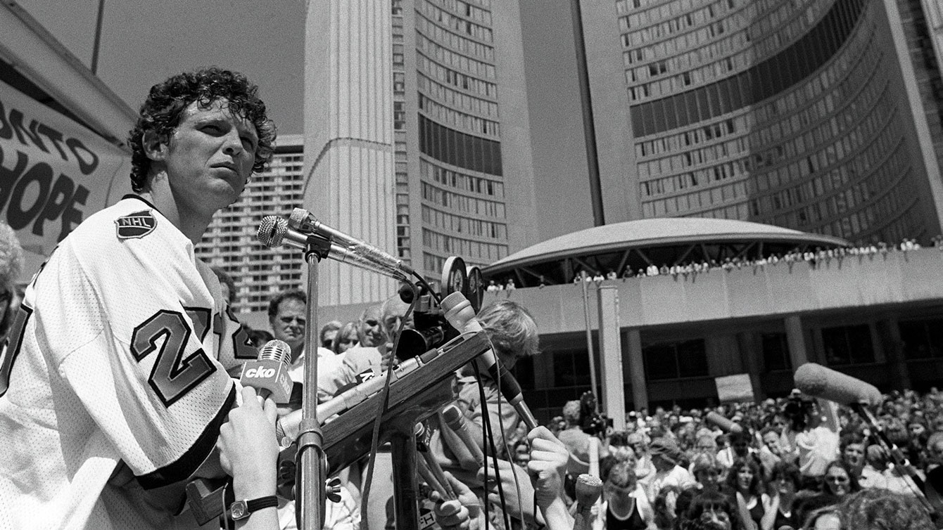 Terry Fox addresses the crowd at Nathan Phillips Square where a reported 10,000 people had gathered to watch him arrive in front of City Hall. Fox is wearing a Darryl Sittler NHL All-Star Game jersey, donated to him by the Toronto Maple Leafs legend who joined Fox on the run up University Avenue.
