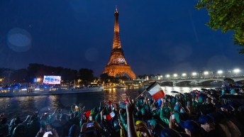 The Eiffel Tower lit up at night with the Olympic rings in white while people wave French flags in the foreground