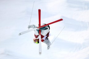 Mikael Kingsbury of Canada competes in the Freestyle Skiing Men's Moguls Qualification 1 event at Phoenix Snow Park in Pyeongchang, South Korea. His knees are bent, and his skis are together as he skis over the right ride of a mogul. His arms are holding his poles as they dig into the snow. He is dressed in a white jacket and snow pants with red Olympic jersey overtop.