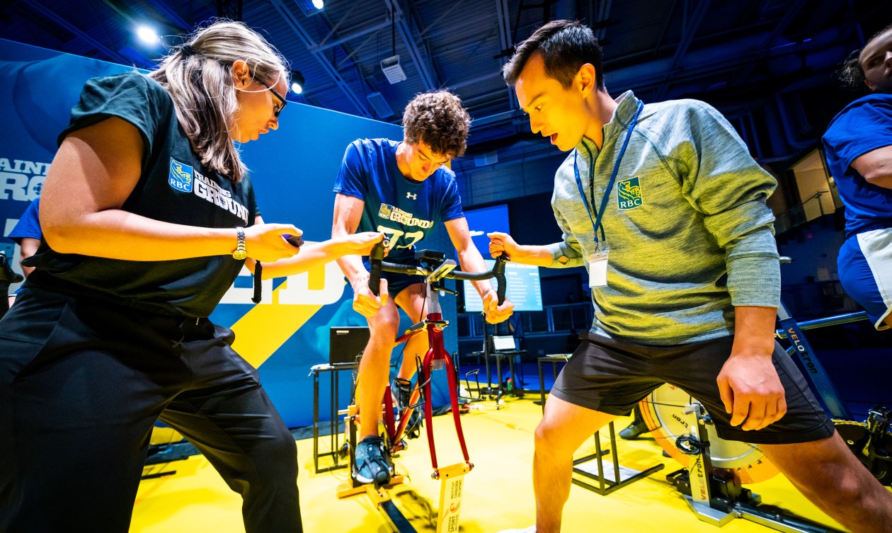 An athlete (centre) rides the sprint bike at the speed station with Patrick Chan (right) cheering him on and a staff (left) keeping track of time.