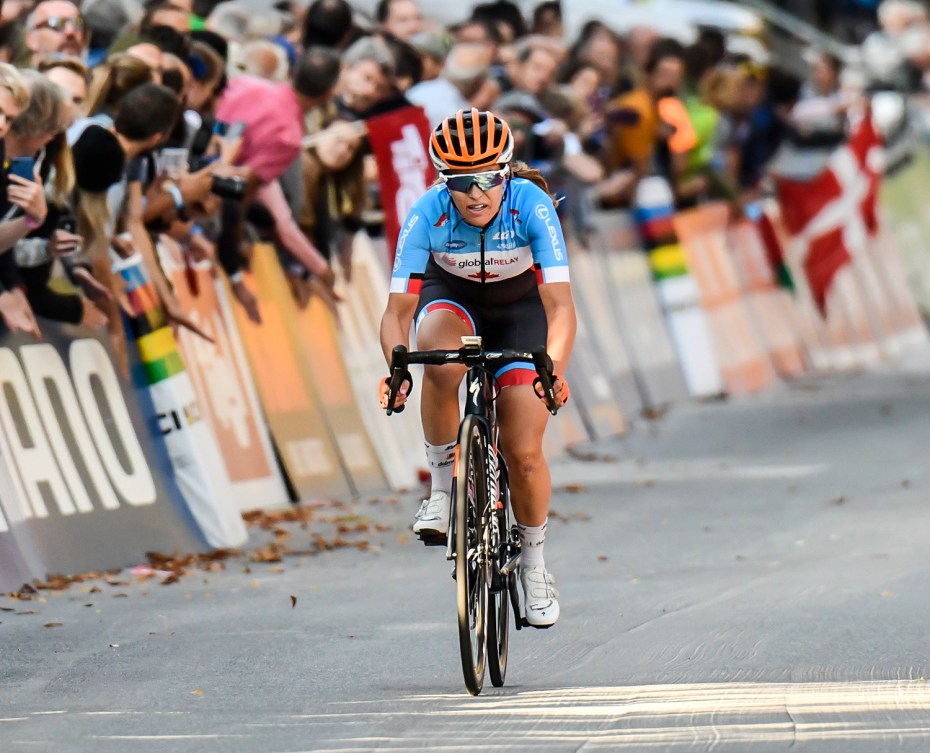 Karol-Ann Canuel cycling at the 2018 UCI World Championships. She is the only cyclist in the shot and is wearing an orange helmet, a blue and white top with black biker shorts that have red and blue detailing.