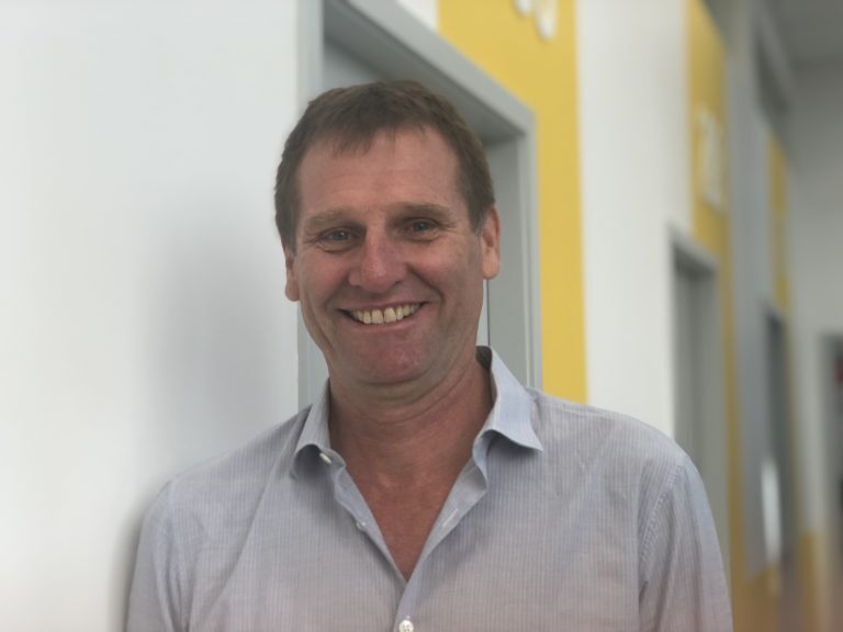 Dr. Mike smiling and standing with his right side against the wall. He is wearing a white collared shirt.