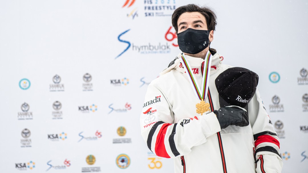 Mikaël Kingsbury holds his black baseball cap over his heart. He is wearing his gold medal around his neck.