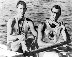 A black and white image of rowers George Hungerford and Roger Jackson sitting in their boat, holding their Olympic medals.