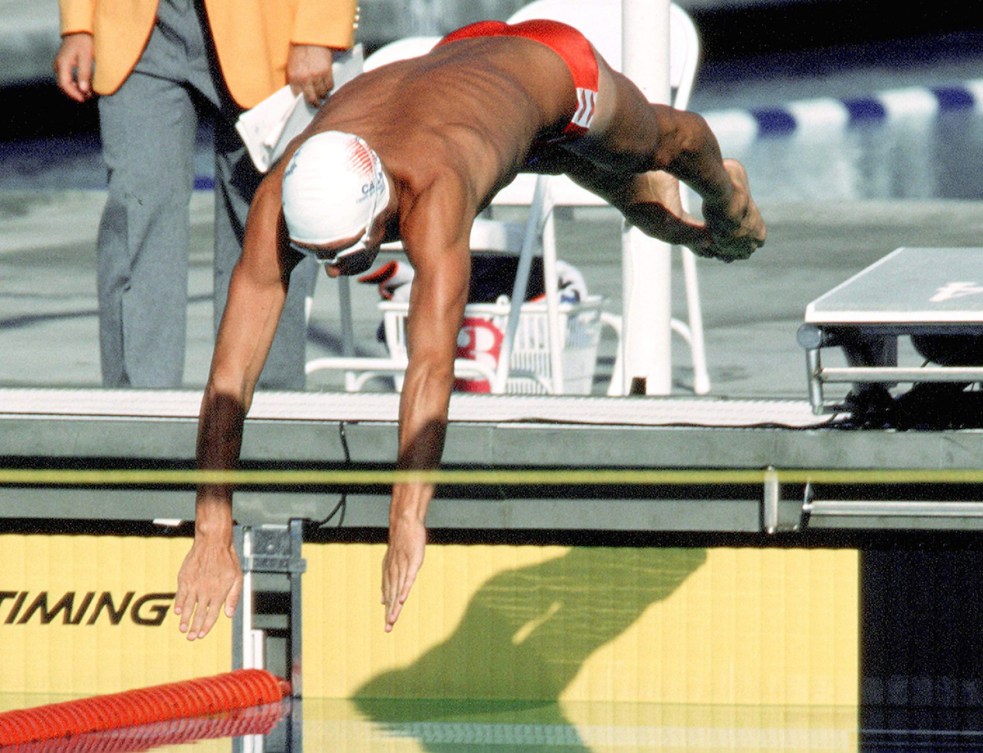 A photo of swimmer Alex Baumann, in a white swim cap, diving into the pool at the 1984 Olympics.
