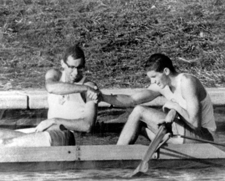 A black and white photo of two men, one wearing glasses, shaking hands while sitting in a boat.