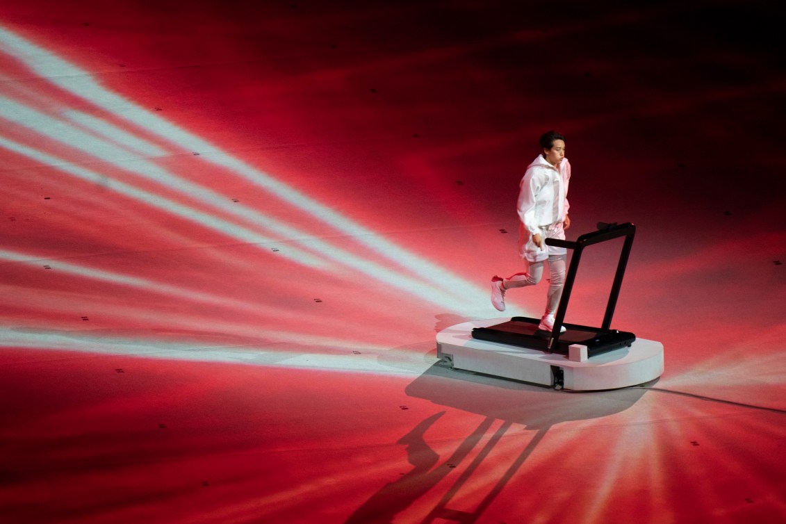 A performer is pictured on a treadmill with beams connecting them to the other performers.
