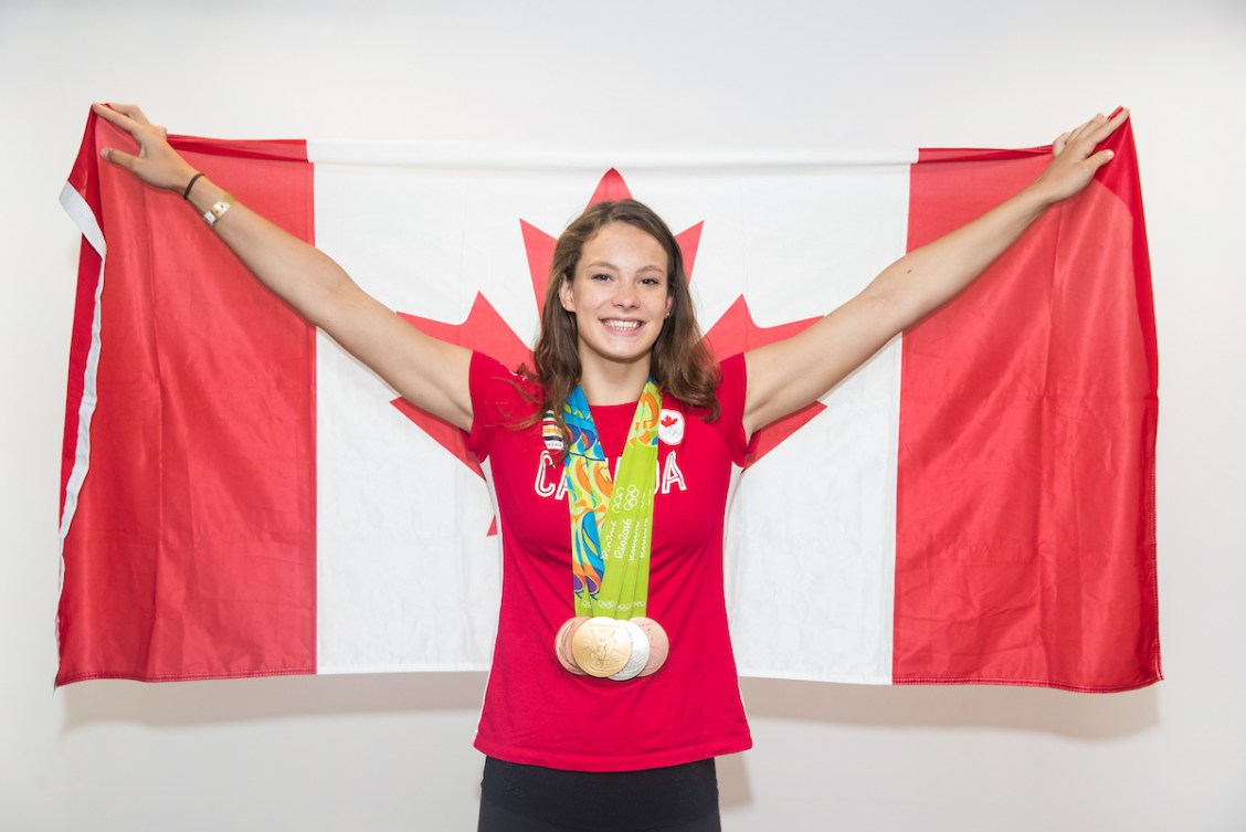 A photo of Penny Oleksiak in a red t-shirt wearing four Olympic medals and holding a Canadian flag.