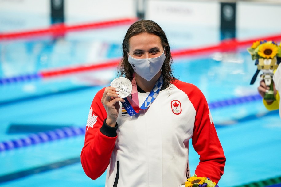 Kylie Masse, wearing a Canada jacket and protective mask, holds up her silver medal in front of the pool.