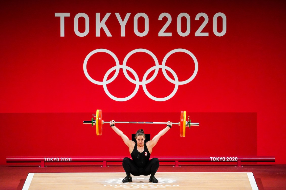 Maude Charron squats with the bar above her head and a large image of the Olympic rings on a wall behind her.