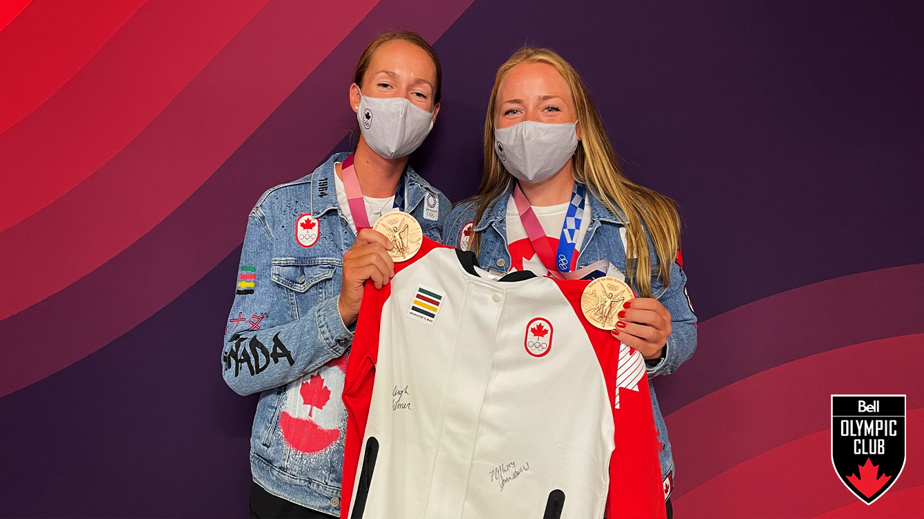 two women wearing masks and jean jackets stand in front of a red backdrop holding a white jacket and two bronze Olympic medals