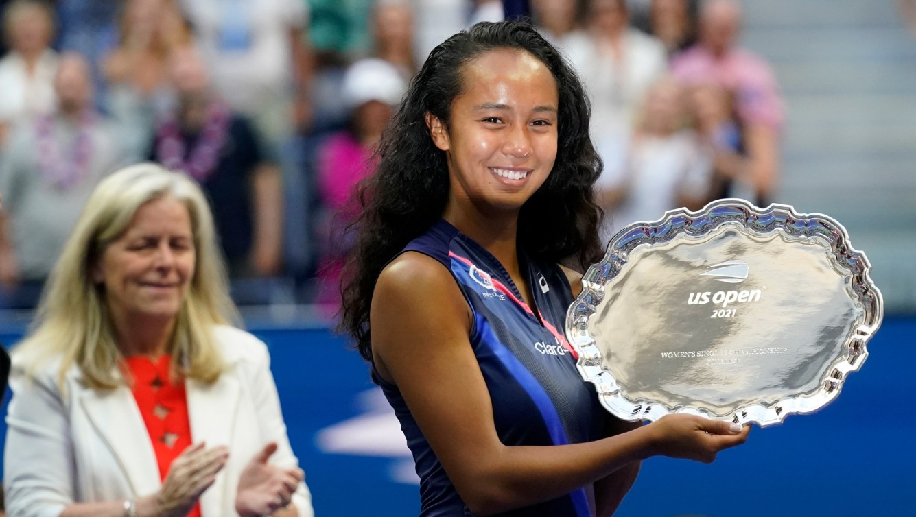 Leylah Fernandez, of Canada, holds up the runner-up trophy (shaped like a silver plate with scalloped edges). The light is reflecting off the plate onto her face.