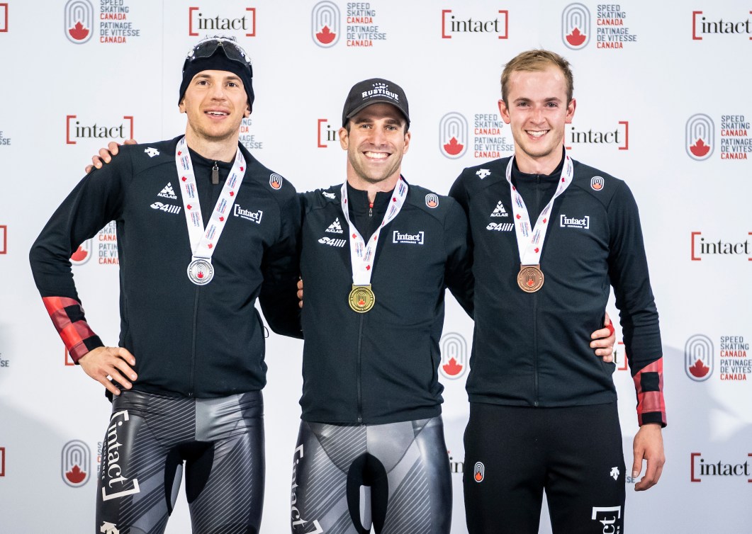 Three male speed skaters stand on a podium 
