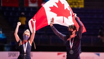 Piper Gilles (left) and Paul Poirier hold up the Canadian flag above their heads. They are wearing their gold medals around their necks.