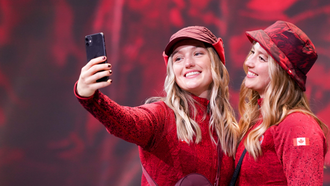 Olympic Freestyle ski athletes and sisters Justine Dufour-Lapointe, left, and Chloe Dufour-Lapointe take a selfie together at the Team Canada x Lululemon Athlete Kit Reveal in Toronto. They are wearing matching red lululemon sweaters and red lululemon hats. They are standing beside each other posed against a red background while Justine’s arm is stretched out to take a photo.