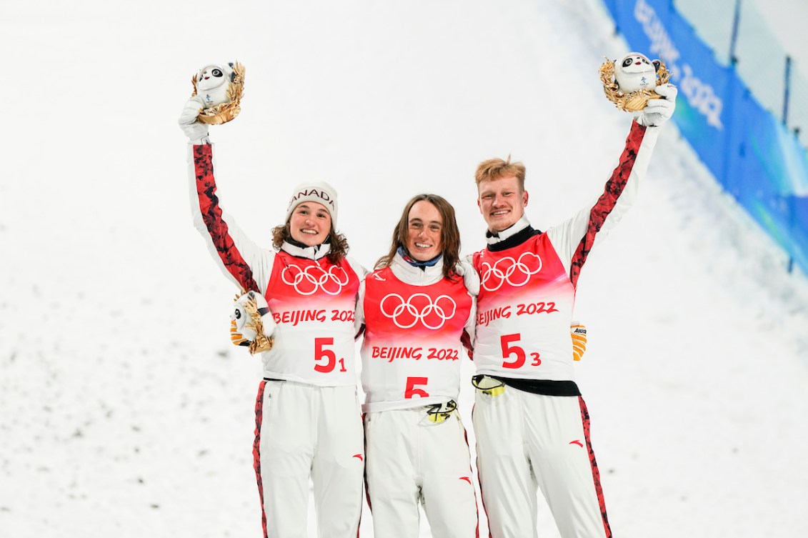 Freestyle skiers stand at the bottom of hill holding up Beijing2022 trophies, and standing arm in arm