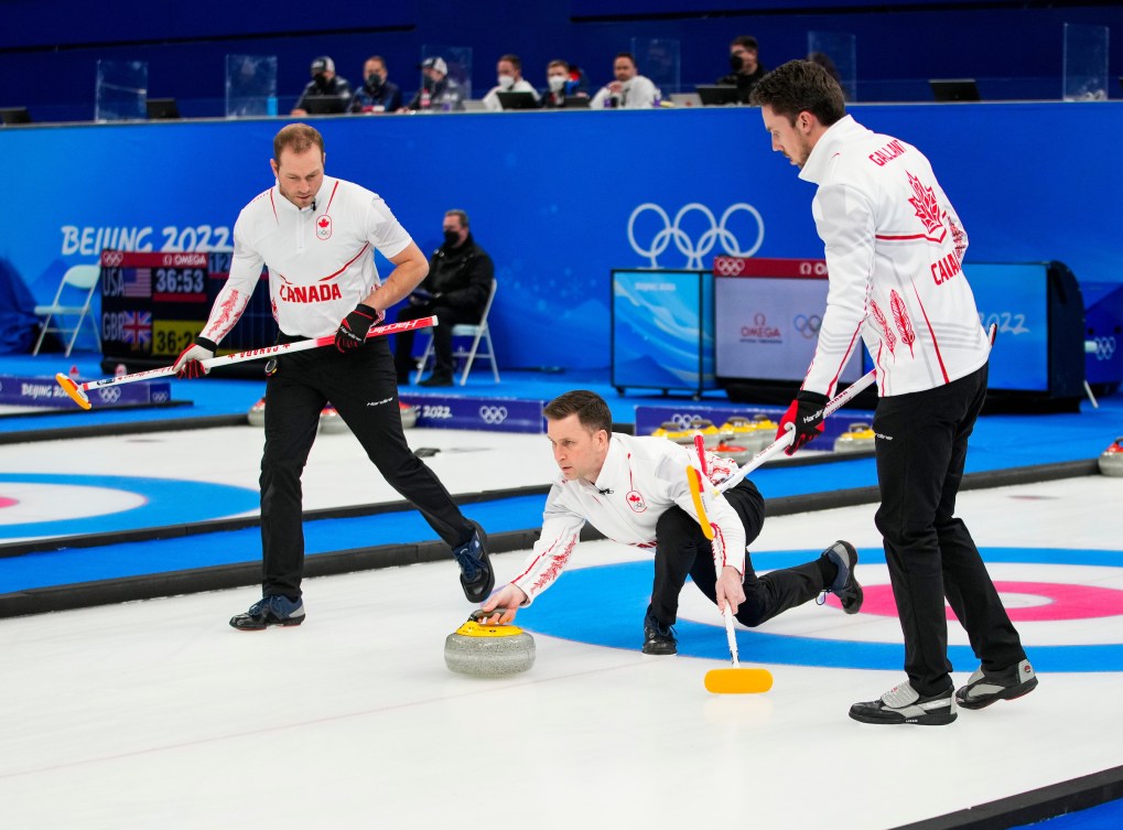 Brad Gushue throws a stone with Geoff Walker and Brett Gallant ready to sweep 