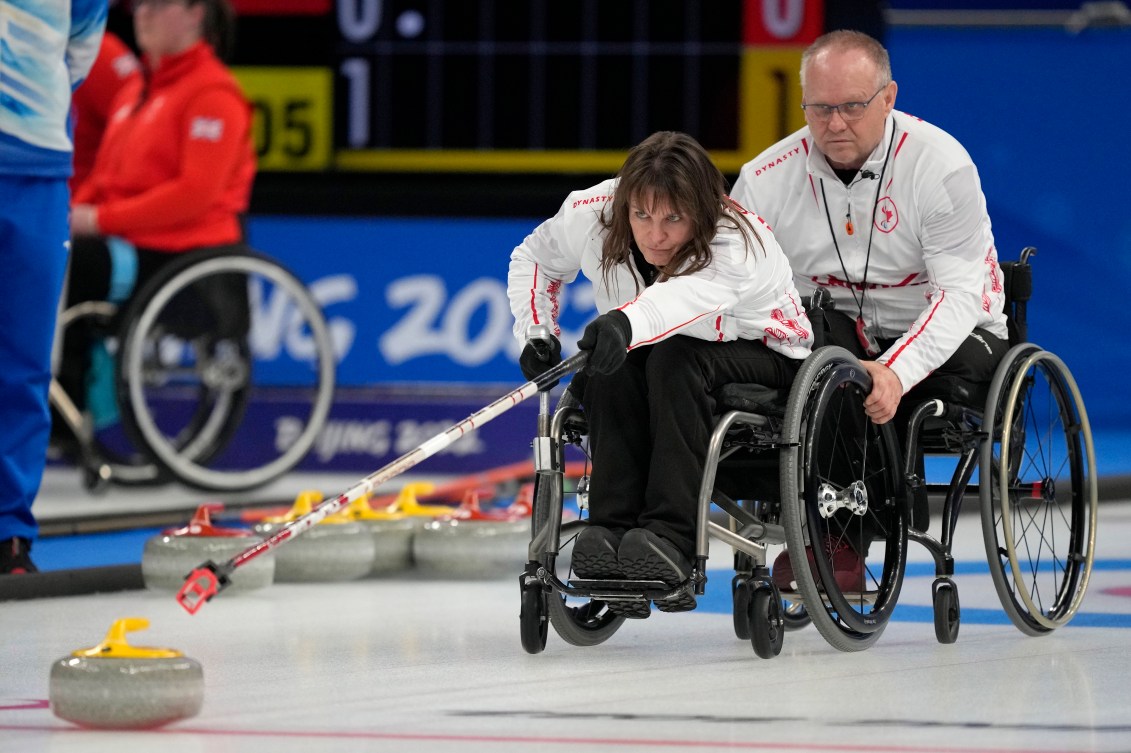One wheelchair curler holds the wheels of a teammate as she pushes the stone with a long stick 
