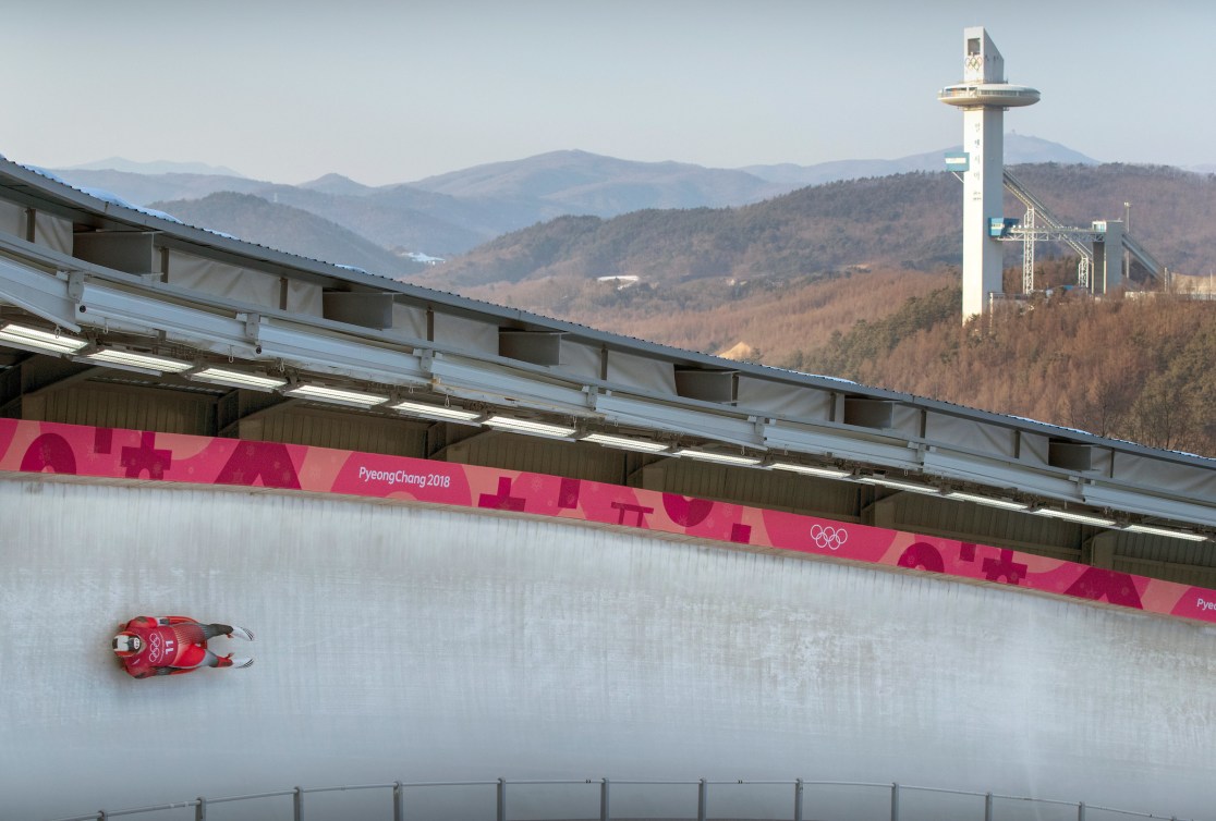 Luger slides on ice track with ski jumping towers in background