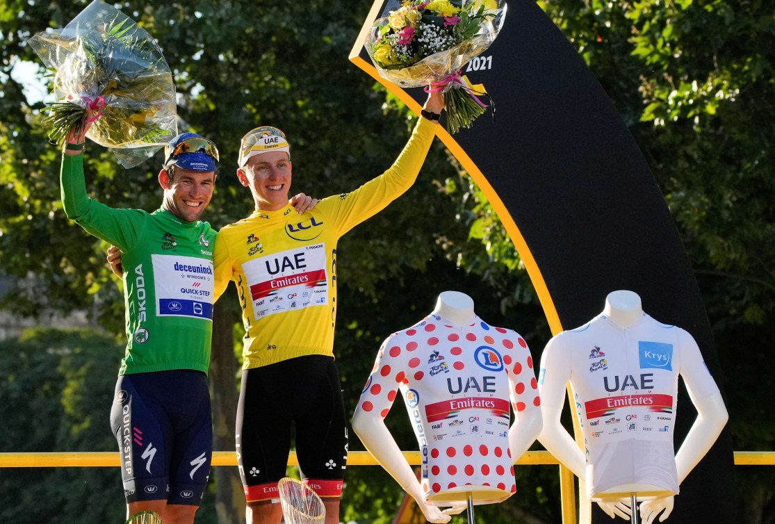 Two cyclists, one in green and one in yellow, stand alongside mannequins wearing a red polka dot jersey and a white jersey