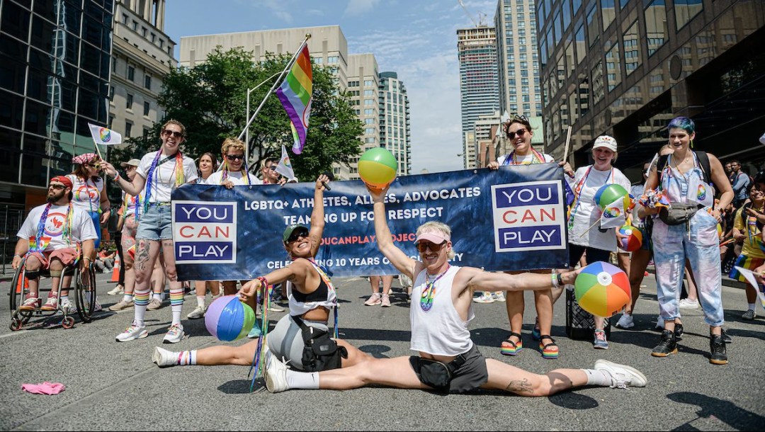 Athletes do splits on the road in front of a You Can Play banner at a Pride Parade