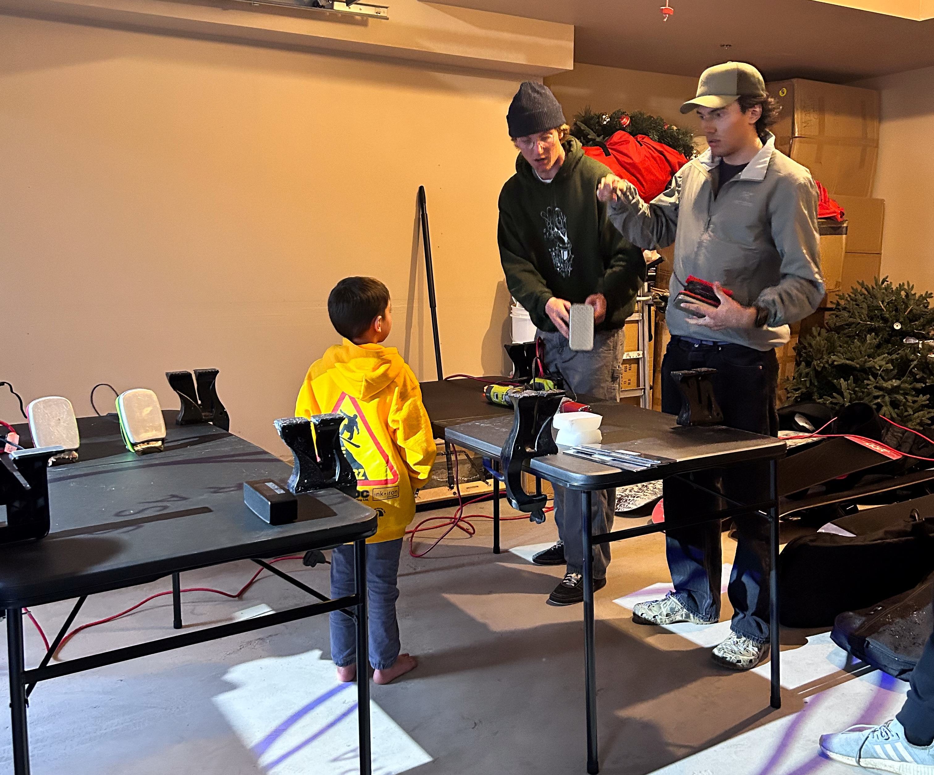 Liam Gill and a friend show a young snowboarder how to wax a snowboard