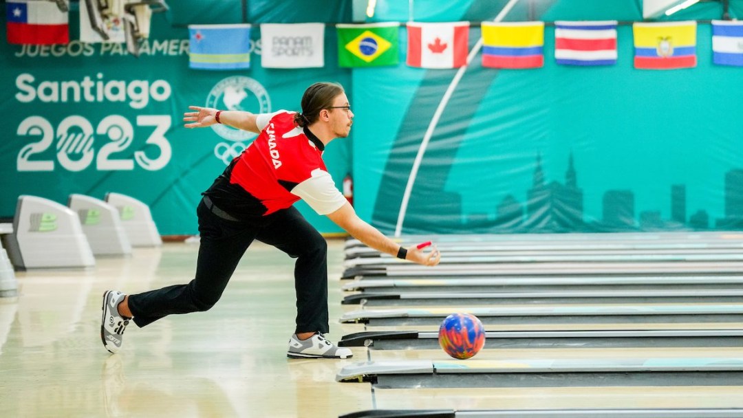 Mitch Hupe in a red shirt and black pants bowls a ball down a lane in an alley