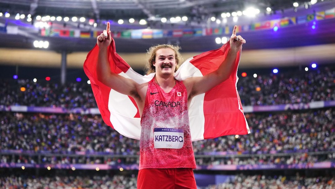 Ethan Katzberg points a finger on each hand to the air as he hold the Canadian flag behind his shoulders