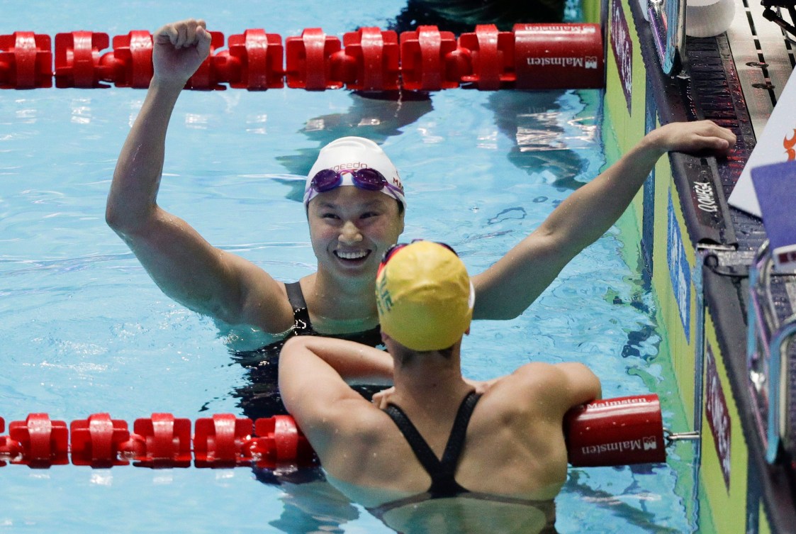 Maggie Mac Neil waves from the water while hanging on to edge of pool 