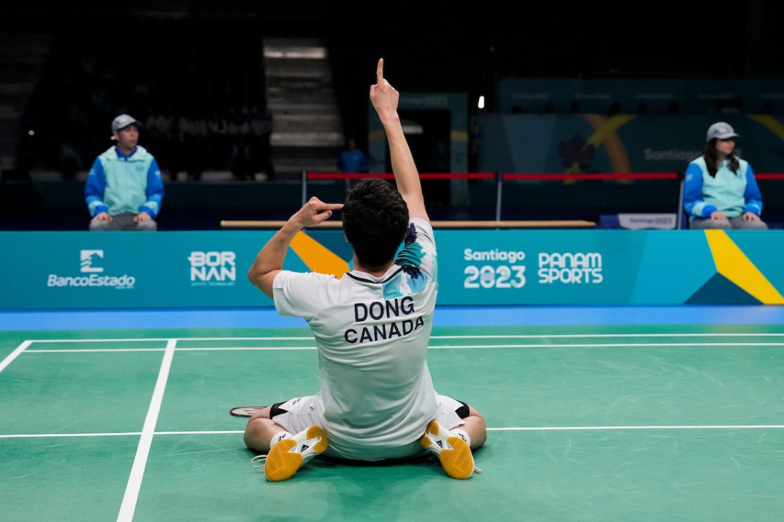 Adam Dong points the one finger on his right hand at the ceiling and one finger of his left hand at his head as he kneels on the court in celebration